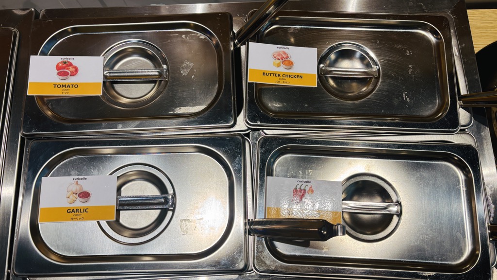 「TOMATO」「BUTTER CHICKEN」「GARLIC」などの札がのった、カレーの入ったステンレスの容器