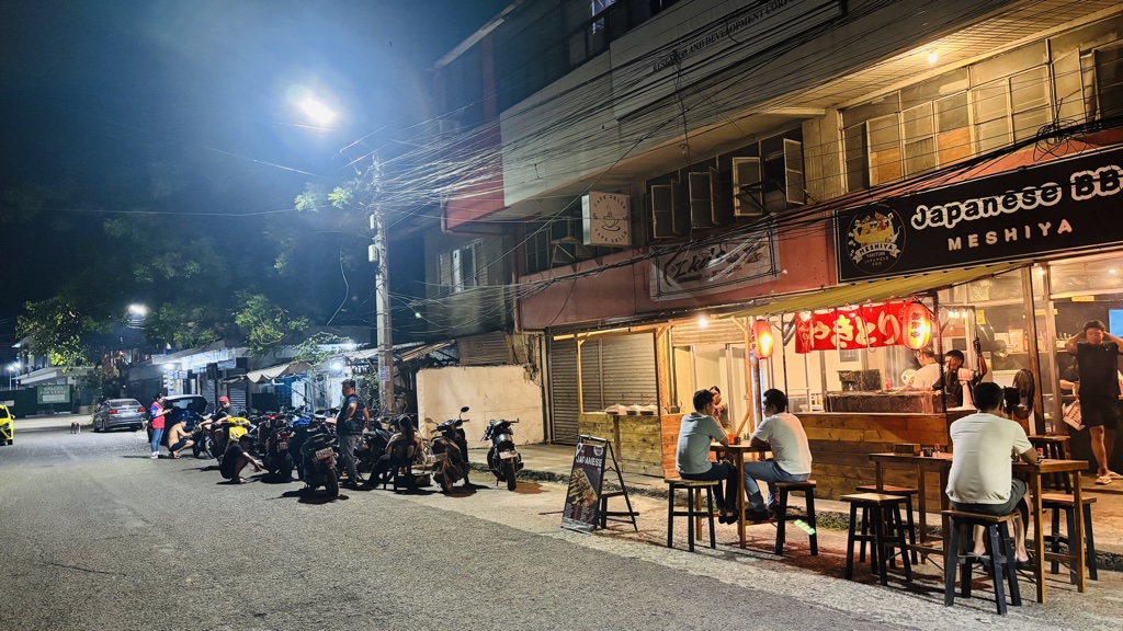 夜のOrlacsan通り。道沿いにバイクが並び、右手にMESHIYAの看板と外のテーブル席