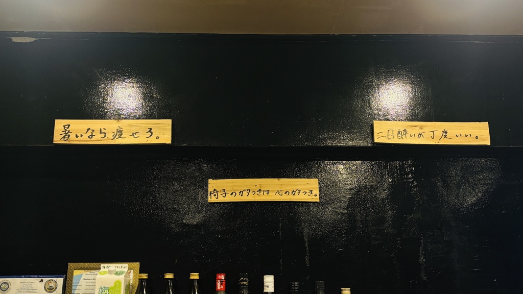 黒い壁に貼られた手書きの木の札。「暑いなら痩せろ。」「二日酔いが丁度いい。」「椅子のガタつきは心のガタつき。」