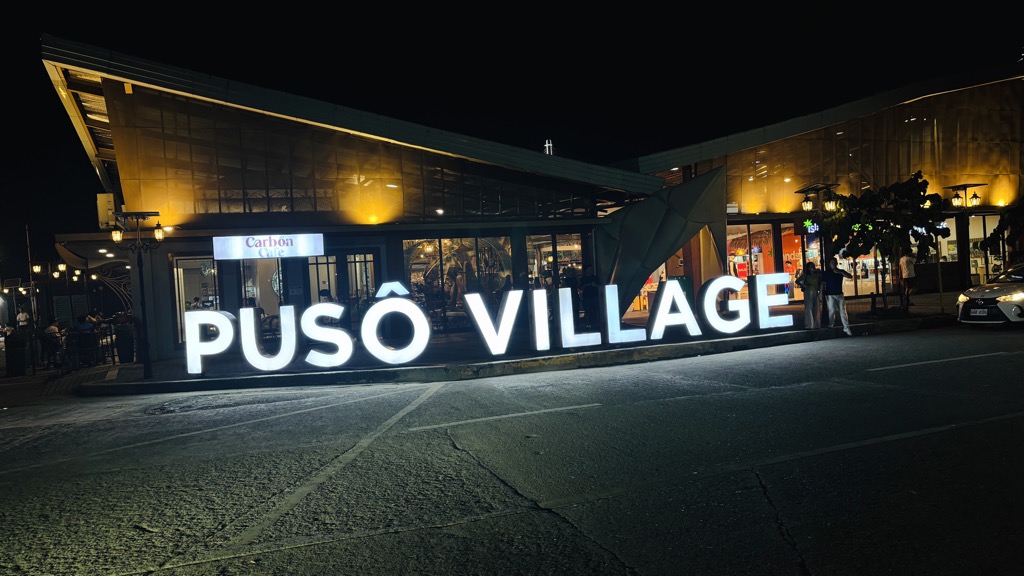 夜にライトアップされた「PUSÔ VILLAGE」の白い文字の看板と建物