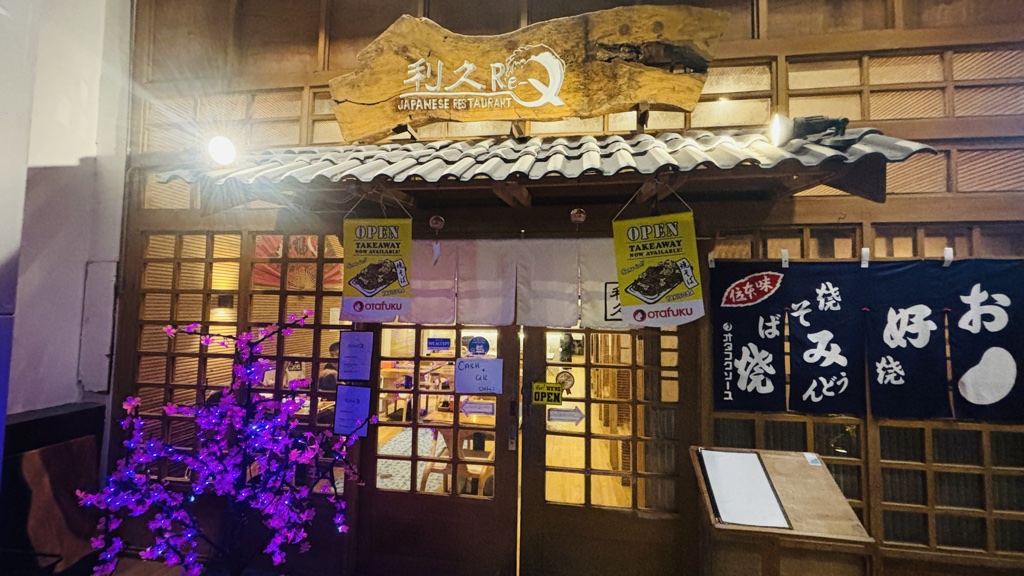 利久（Re-Q）の外観。木の看板と瓦屋根、白いのれん、右側にお好み焼ののれん、左に紫色に光る桜の木